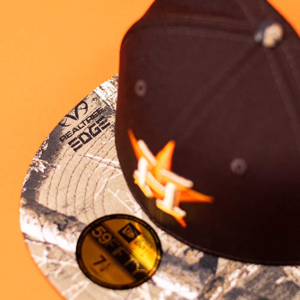 A close-up of the Old Head RTC Wood Oil Houston Astros New Era Hat 5950 Fitted, featuring a black and camo design, Astros star logo, gold 59FIFTY sticker on the camo flat brim, set against an orange background.