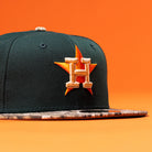 The Greenhand RTC Oil Houston Astros New Era Hat 5950 Fitted features a dark green color, a camouflage brim, and an embroidered orange and gold H with a star on the front, shown against an orange background.