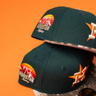Two Greenhand RTC Oil Houston Astros New Era 5950 Fitted hats in dark green, featuring orange/yellow Houston and oil pump embroidery with an H star logo on the side, are shown on an orange surface—ideal for Astros fans.