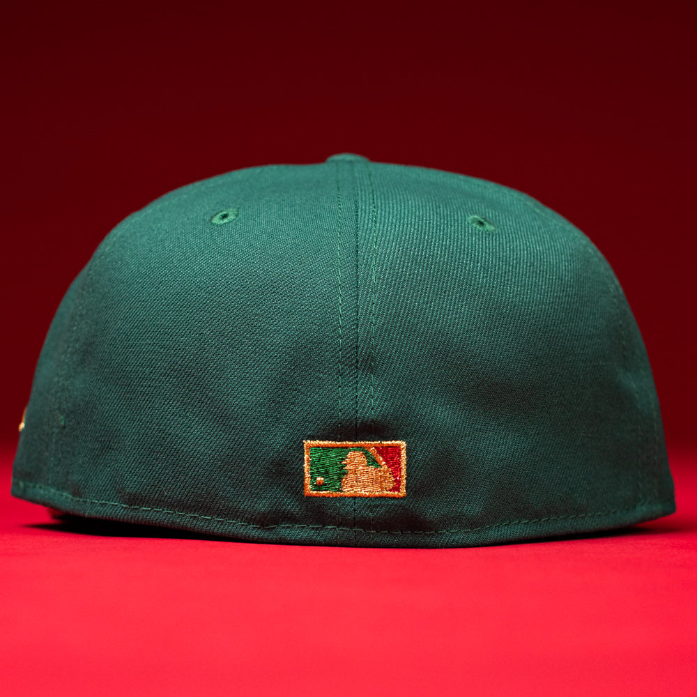 Against a vibrant red backdrop, the La Virgencita Green Red Gold New Era Houston Astros Hat 5950 Fitted is shown from the back, highlighting a small embroidered Major League Baseball logo in gold, green, and white near the bottom edge—a subtle tribute to Mexican culture that lends this cap its unique and meaningful appeal.
