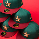 A stack of La Virgencita Green Red Gold New Era Houston Astros Hat 5950 Fitted baseball caps, featuring gold star logos and New Era stickers on the visors, inspired by designs that celebrate Mexican culture, set against a bright red background.