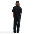 A person with long curly hair, wearing the HV2195010 Max 90 Skate T-Shirt SB Tee Black from Nike SB Skateboarding, black pants, and a dark cap stands facing away from the camera against a plain white background.
