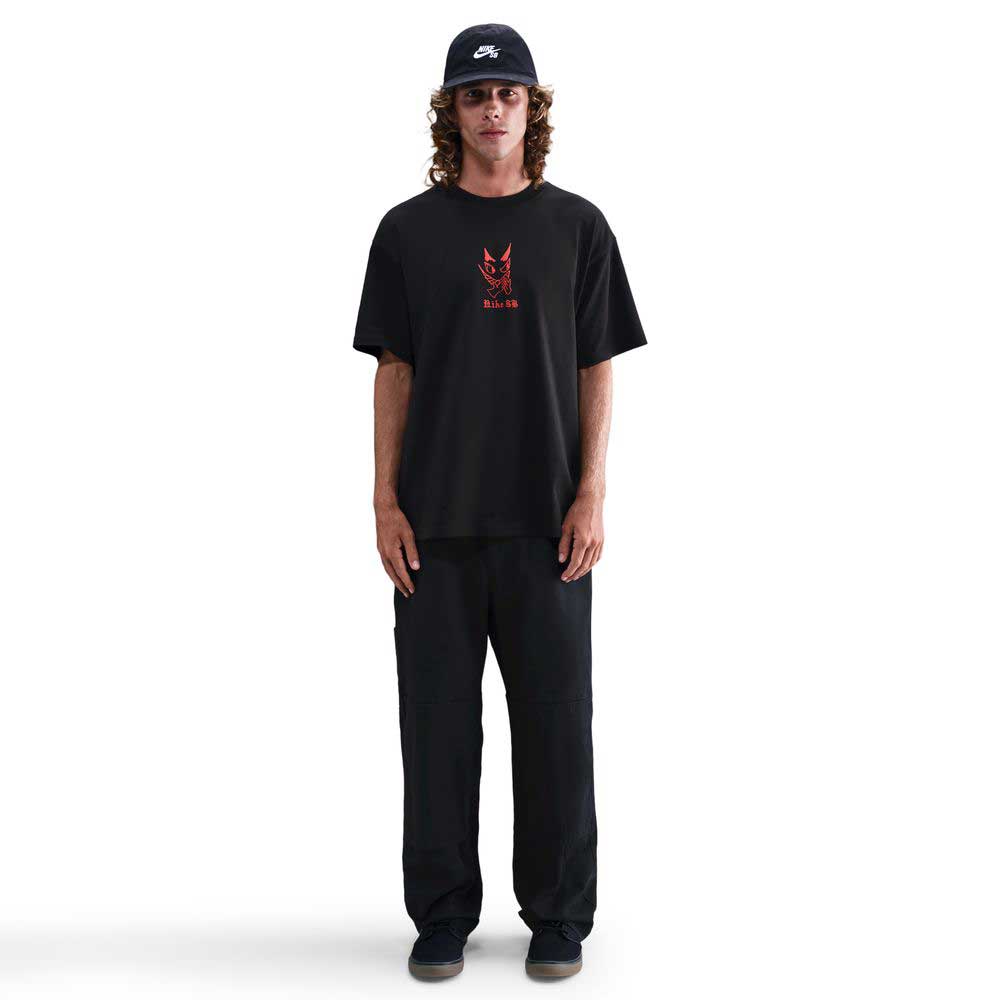 A person with long hair, wearing a black cap, black pants, dark shoes, and the Nike SB Max 90 Skate T-Shirt (black with a red graphic), stands facing forward against a plain white background.