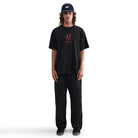 A person with long hair, wearing a black cap, black pants, dark shoes, and the Nike SB Max 90 Skate T-Shirt (black with a red graphic), stands facing forward against a plain white background.