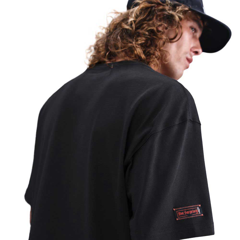 A person with long curly hair, seen from the back and looking over their shoulder, wears the Nike SB Max 90 Skate T-Shirt in black—a midweight cotton tee with a small rectangular patch on the sleeve, designed for durable daily comfort.