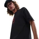 A young man with long curly hair, wearing the Nike SB Max 90 Skate T-Shirt (HV2195010) and a black cap, looks down at the camera against a plain white background.