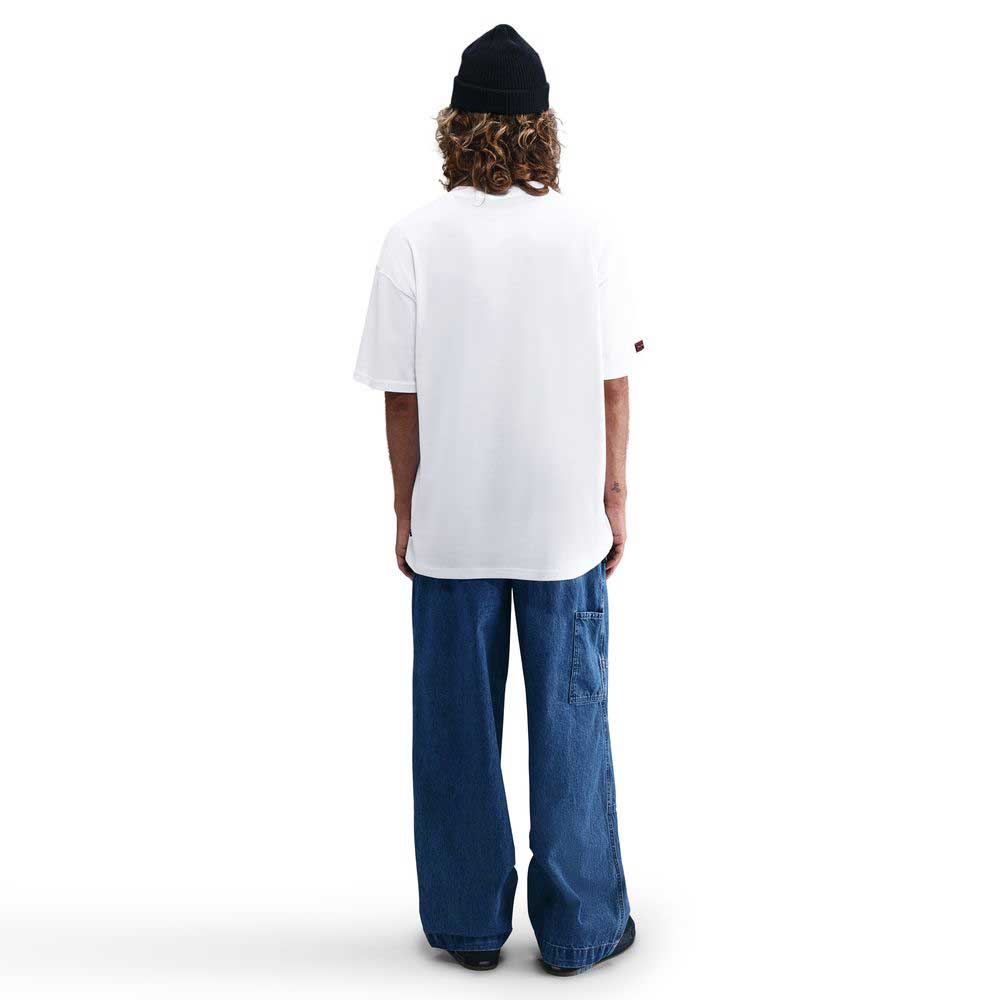A person with curly hair, wearing a black beanie and Nike SB Max 90 Skate T-Shirt SB Tee in white with wide blue jeans, stands facing away against a plain white background.