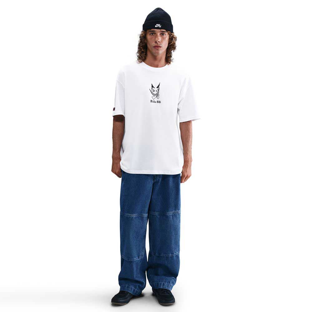 A person with long curly hair wears a black beanie, wide-leg blue jeans, black shoes, and the Nike SB Max 90 Skate T-Shirt in white (HV2195100), standing against a plain white background for durable daily comfort.