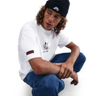 A person with curly hair wears a black Nike beanie, blue jeans, and the HV2195100 Max 90 Skate T-Shirt SB Tee—a midweight, white Nike SB skateboarding tee with screenprinted graphics—against a plain white background for daily comfort.