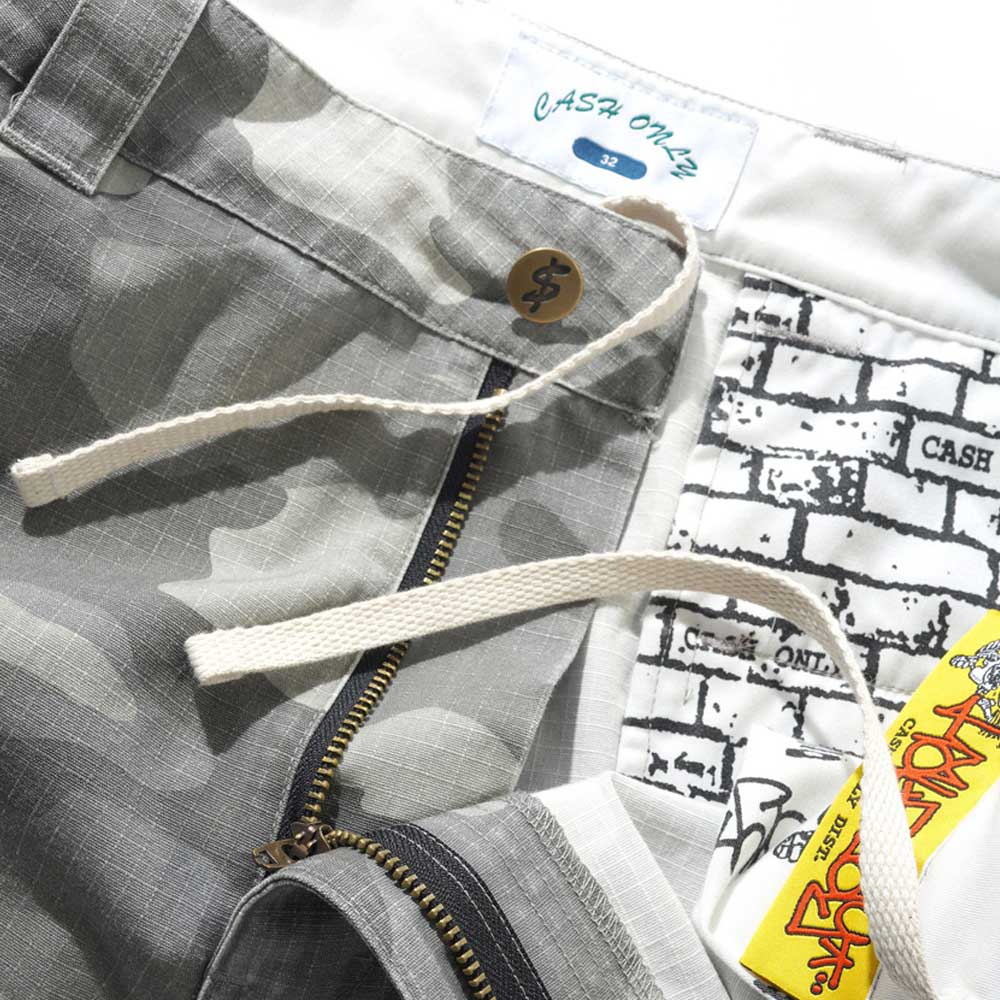 A close-up of Halfway Shorts Grey Camouflage Ripstop Cash Only COD152601 shows baggy cotton twill with an adjustable drawcord, brass button, zipper, brick-patterned inner waistband with “CASH ONLY” branding, and a yellow tag with red text.