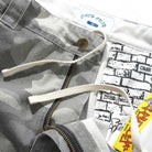 A close-up of Halfway Shorts Grey Camouflage Ripstop Cash Only COD152601 shows baggy cotton twill with an adjustable drawcord, brass button, zipper, brick-patterned inner waistband with “CASH ONLY” branding, and a yellow tag with red text.
