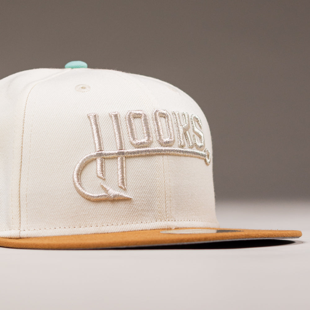 The Hookset Corpus Christi Hooks New Era Hat 5950 Fitted is a cream-colored cap with "HOOKS" embroidered in white on the front and a tan brim, inspired by the Corpus Christi Hooks, shown on a neutral background.