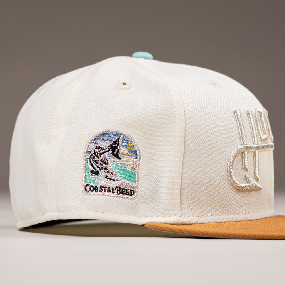 The Hookset Corpus Christi Hooks New Era Hat 5950 Fitted is a white baseball cap with a tan brim, featuring an embroidered black and white dog patch on a beach, COASTAL BRED text, and bold white raised front lettering.