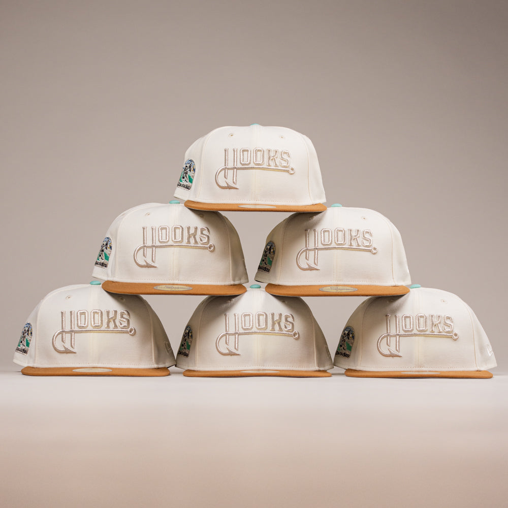 Six white The Hookset Corpus Christi Hooks New Era Hat 5950 Fitted caps with gold brims and gold "LOOKS" embroidery are stacked pyramid-style, featuring a vibrant side patch honoring the Hooks on the top and side hats.