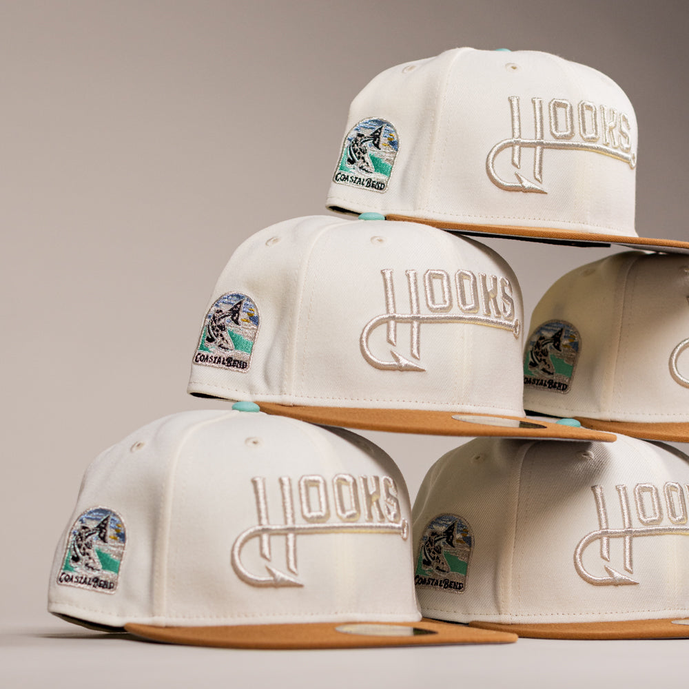 Six cream-colored The Hookset Corpus Christi Hooks New Era Hat 5950 Fitted caps are stacked in a pyramid, each with a bold Hooks logo featuring a hook on the front and a colorful angler fishing patch on the side.