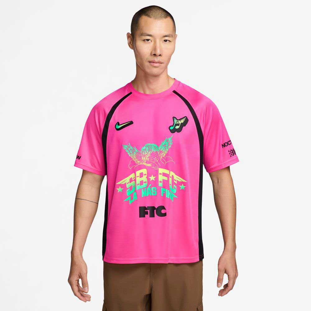 A man stands against a white background wearing a Hyper Pink Black IR6760645 Skate Jersey by Nike SB Skateboarding, featuring neon green and blue graphics, black side panels, multiple logos, stylized text, and an easy-care polyester body.