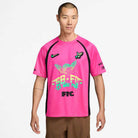 A man stands against a white background wearing a Hyper Pink Black IR6760645 Skate Jersey by Nike SB Skateboarding, featuring neon green and blue graphics, black side panels, multiple logos, stylized text, and an easy-care polyester body.