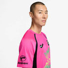 A man with short hair wears the Hyper Pink Black IR6760645 Skate Jersey from Nike SB Skateboarding, featuring black accents, a palm tree graphic, "Andrew" and a sleeve logo. The comfy polyester jersey is machine washable. He stands against a white background.