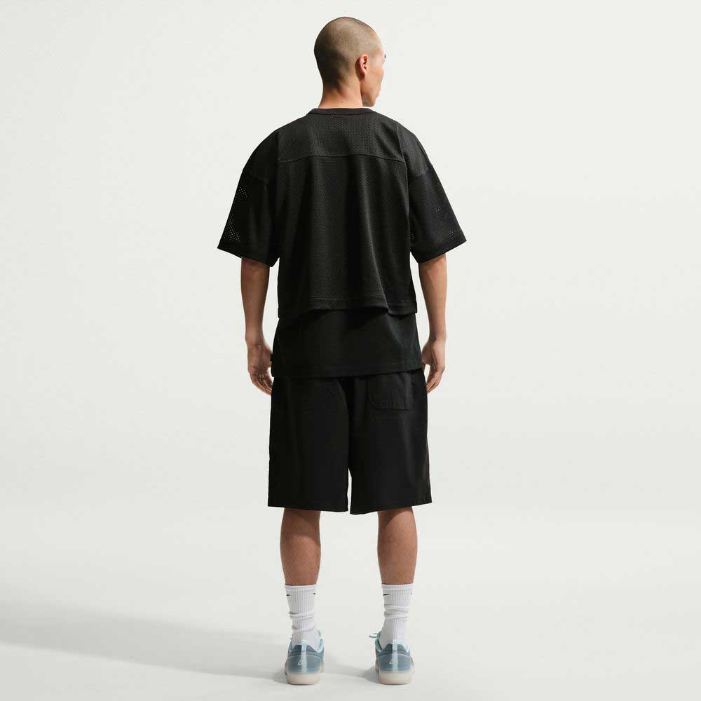 A person with a shaved head faces away, wearing a loose black mesh shirt, IF1096010 Black White Skate Shorts Nike SB, white socks, and light-colored sneakers against a plain light background.
