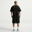 A person with a shaved head faces away, wearing a loose black mesh shirt, IF1096010 Black White Skate Shorts Nike SB, white socks, and light-colored sneakers against a plain light background.