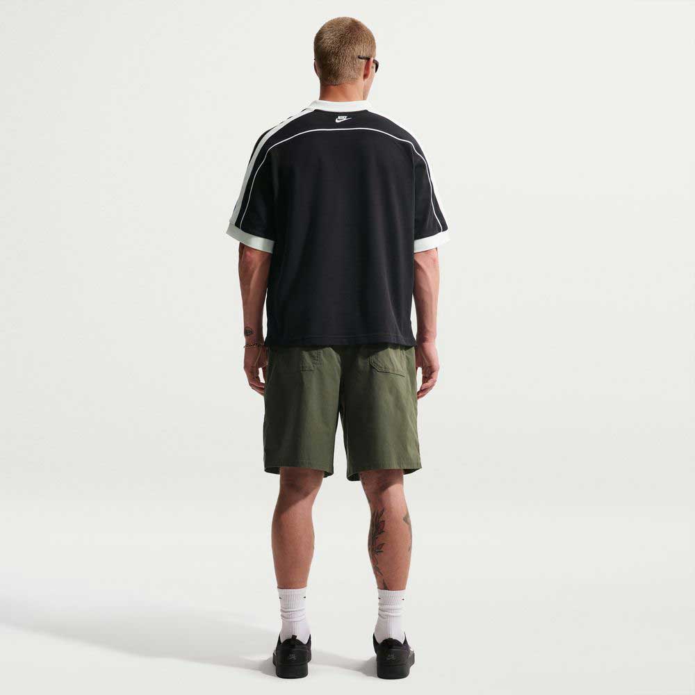 A person with short hair faces away in skate style: a black Nike shirt with white trim, IF1096325 Cargo Khaki White Skate Shorts Nike SB, white socks, and black shoes against a plain white background.