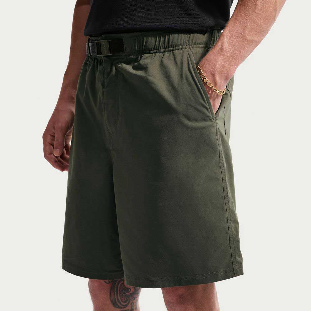 Wearing IF1096325 Cargo Khaki White Skate Shorts by Nike SB, a person stands against a plain light background with hands in the pockets, paired with a black belt, black shirt, and gold chain bracelet for a skate-ready look.