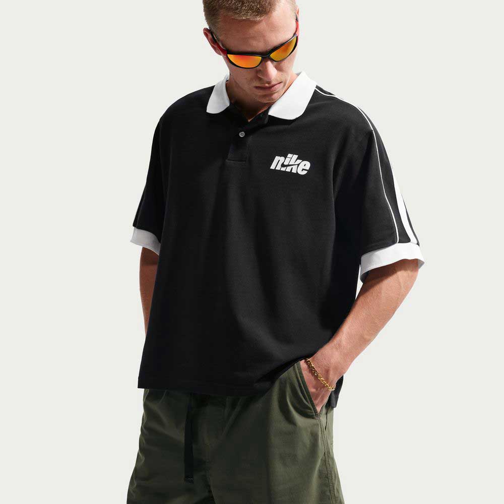A man in sunglasses, a black Nike polo with white trim and logo, and IF1096325 Cargo Khaki White Skate Shorts from Nike SB—made with sweat-wicking fabric—stands with one hand in his pocket, looking down.