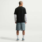 A man faces away, wearing a black short-sleeve shirt over a white long-sleeve, IF1096382 Mineral Slate Blue Skate Shorts Nike SB, white socks, and light blue sneakers, all set against a plain white background.