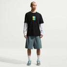 A person stands against a plain background wearing a black T-shirt with a colorful graphic, a long white shirt underneath, IF1096382 Mineral Slate Blue Skate Shorts Nike SB, white socks, and teal sneakers—a skate-ready fit.