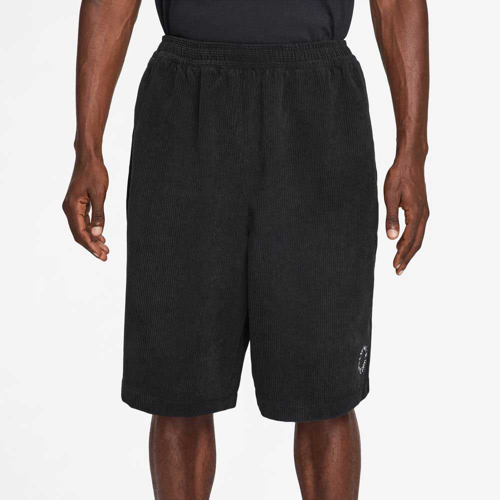 A person wearing II4426010 Black White Corduroy Skate Shorts Nike SB and a black shirt stands with arms relaxed by their sides against a plain background.