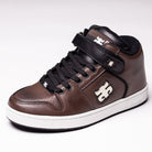 Grasshopper Brown Leather I Path Shoes Skateboarding in brown and black feature a high-top design crafted from Grasshopper brown leather, with a classic white sole, black laces, and a Velcro strap. The shoes showcase a distinctive white puzzle piece logo on the side and tongue and are fitted with an Ortholite insole for enhanced comfort. Photographed against a plain white background.