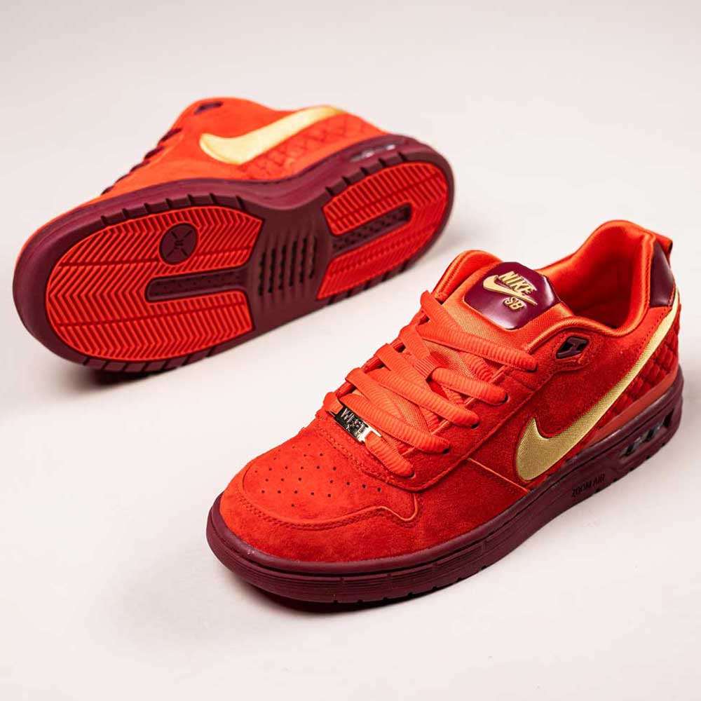 A pair of vibrant IQ5648600 Habanero Red Club Gold Team Red Paul Rodriguez Zoom Air Low Nike SB suede skateboarding shoes with gold swooshes, burgundy soles, and red laces. One shoe stands upright; the other lies on its side to display the sole.