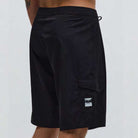 A person is photographed from the back wearing the Crux 20 inch Trunk Black Lime Former—loose-fitting black shorts featuring a side pocket and patch. Part of their lower back and a tattoo on their upper arm are visible.