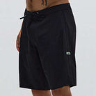 A person wearing the Crux 20 inch Trunk Black Lime Former shorts, featuring a drawstring waist, stands against a light gray background. Only the lower torso and upper legs are visible.