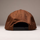 The Truck Co Legacy Brown Hat by Independent Skateboards is a tan, unstructured snapback with a black adjustable strap and low-profile fit, shown from the back on a neutral background. Unisex, OS.