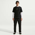 A person stands facing forward against a plain white background, wearing black pants, black sneakers, headphones, a long-sleeve white shirt, and the Ishod Tee Black IH1437010 Nike SB T-Shirt.