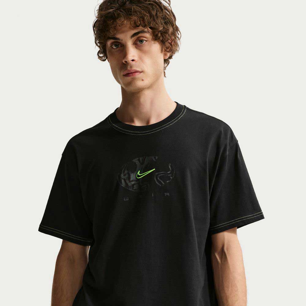 A young man with curly brown hair wears the Ishod Tee Black IH1437010 Nike SB T-Shirt, made from midweight cotton with a neon green logo and subtle graphic, standing against a plain white background and gazing slightly to the side.