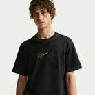 A young man with curly brown hair wears the Ishod Tee Black IH1437010 Nike SB T-Shirt, made from midweight cotton with a neon green logo and subtle graphic, standing against a plain white background and gazing slightly to the side.
