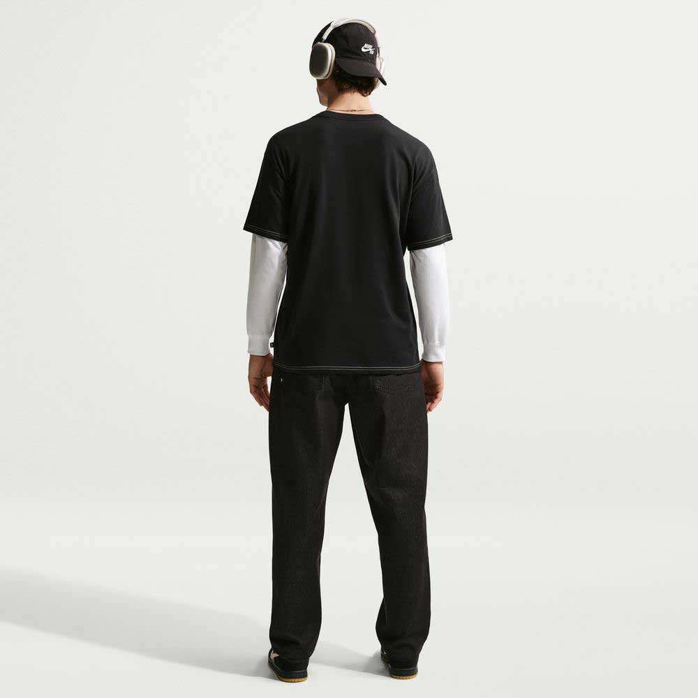 A person faces away from the camera, wearing the Ishod Tee Black IH1437010 Nike SB T-Shirt over a long-sleeve white shirt, with headphones, a black cap, and dark pants against a plain white background.