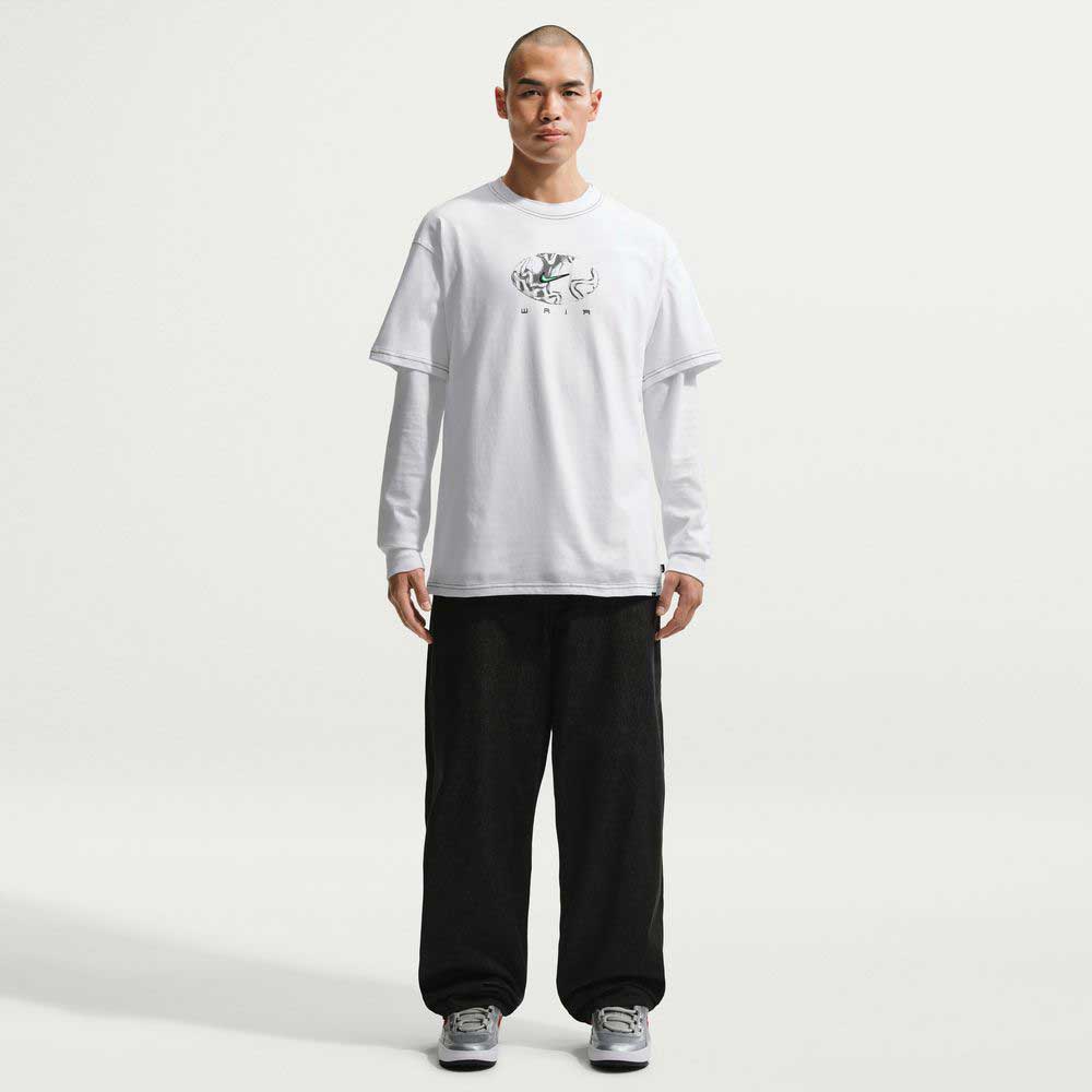 A person stands facing forward in a white Ishod Tee White IH1437100 Nike SB T-Shirt over a long-sleeve shirt, loose black pants, and gray sneakers, set against a plain white background.