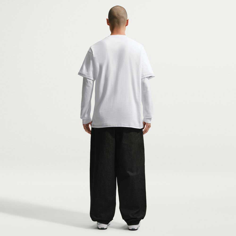 A person stands with their back to the camera, wearing the Ishod Tee White IH1437100 Nike SB T-Shirt over a long-sleeve shirt, loose black pants, and white sneakers against a plain light background.