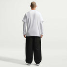 A person stands with their back to the camera, wearing the Ishod Tee White IH1437100 Nike SB T-Shirt over a long-sleeve shirt, loose black pants, and white sneakers against a plain light background.