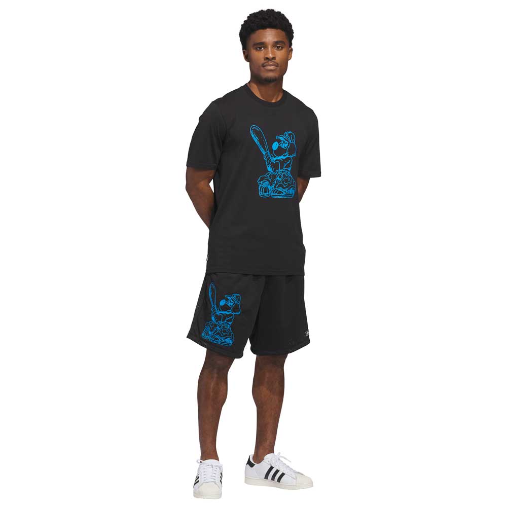 A man stands in skate style, wearing KC6308 Kogan Shorts Blue Black Adidas Skateboarding and a black T-shirt with a blue cartoon graphic. He completes the look with white adidas Originals sneakers featuring bold black stripes.