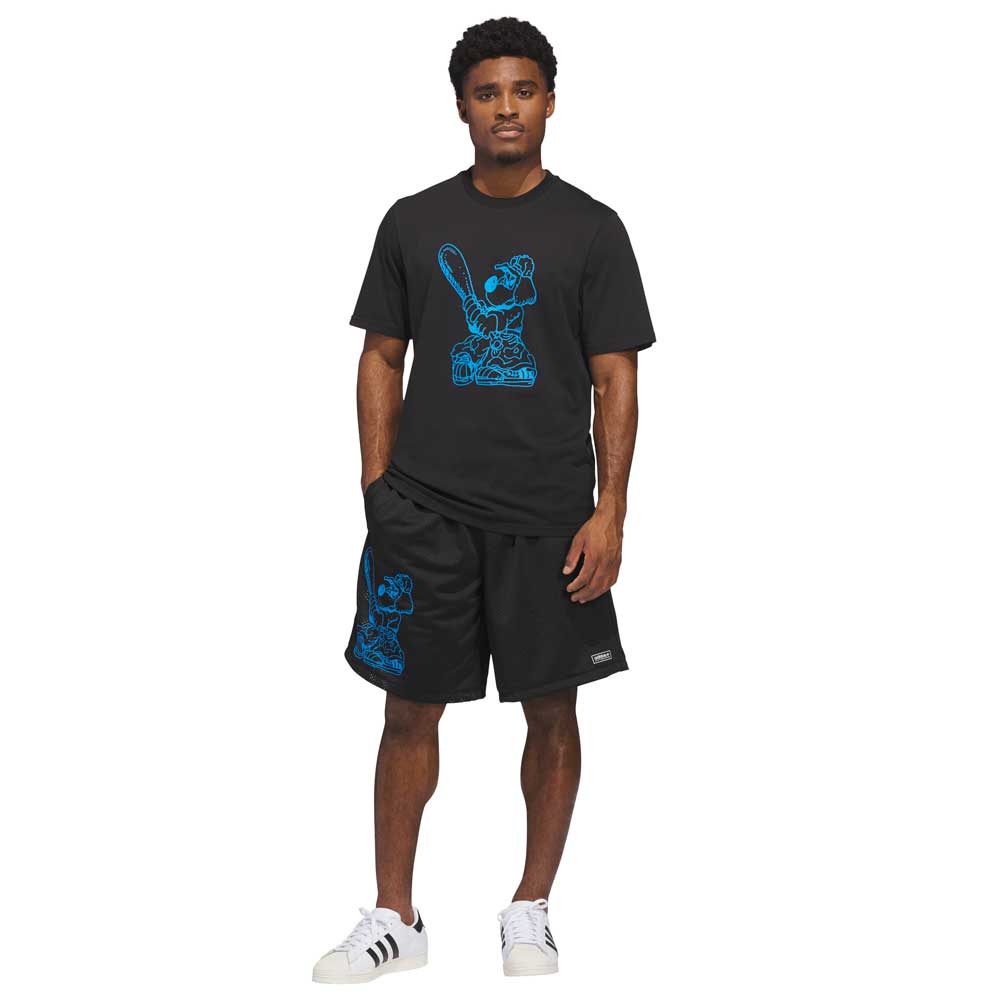 A man stands facing forward, wearing a black Kogancult T-shirt and KC6308 Kogan Shorts Blue Black Adidas Skateboarding, featuring blue and black graphics. He has one hand in his pocket and sports white Adidas sneakers with black stripes.