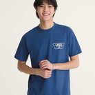 A person with straight black hair smiles while wearing the Knu Patch VN000WU85TU1 True Navy Blue Short Sleeve Tee from Vans Skateboarding, featuring a white logo on the left chest, set against a plain, light background.