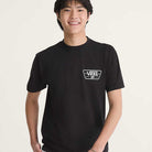 A smiling person with short dark hair wears a Knu Patch VN000WU8BLK1 Black Short Sleeve Tee by Vans Skateboarding, featuring a white Vans logo on the left chest, against a plain light background.