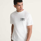 Wearing the Knu Patch VN000WU8WHT1 White Short Sleeve Tee from Vans Skateboarding, featuring a small Vans logo on the left chest, a person stands against a neutral background looking to the side.