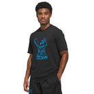 A man in the Kogan Dog Tee Black Blue KC6304 by Adidas Skateboarding stands with one hand in his pocket, its bold blue graphic on black channeling effortless skate culture. He completes the look with black shorts.