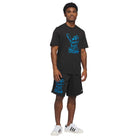 A man stands facing forward, wearing the Kogan Dog Tee Black Blue KC6304 Adidas Skateboarding and black shorts with blue bear graphics, paired with white Adidas sneakers with black stripes.