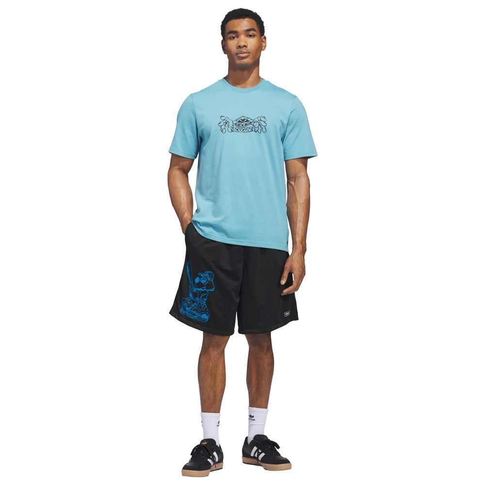 A man stands against a white background wearing the Kogan Logo Tee Blue Black KC6301 Adidas Skateboarding t-shirt, black shorts with blue details, white socks, and black adidas sneakers with white stripes, facing forward with one hand in his pocket.